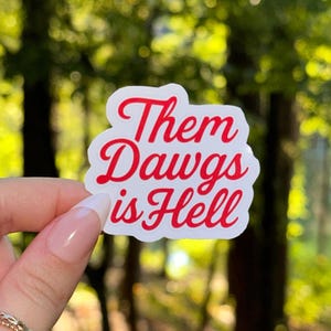 May include: A white sticker with a red border and the text "Them Dawgs is Hell" in a cursive font. The sticker is held up against a blurred green and brown background of trees.