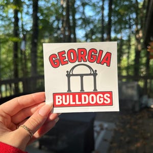 May include: A white sticker with the word "GEORGIA" in red arched letters above a black illustration of a building with three columns. The text "BULLDOGS" is in a red rectangle below.