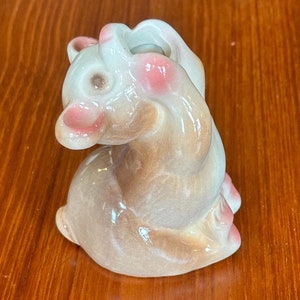 May include: A ceramic bunny figurine in a seated position, with a glossy finish. The bunny has brown and white fur, with pink accents on the ears and nose. The figurine is a decorative item.