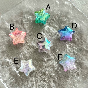 May include: Six iridescent star-shaped embellishments in various colors, including green, pink, blue, and purple. The stars are arranged in a circle on a white background.