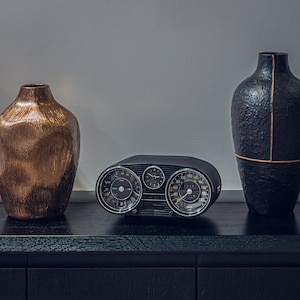 Puede incluir: Un tablero de instrumentos de coche vintage negro con tres diales redondos, incluyendo un velocímetro, un tacómetro y un reloj. El tablero está sentado sobre una superficie de madera negra con dos jarrones decorativos, uno dorado y otro negro.