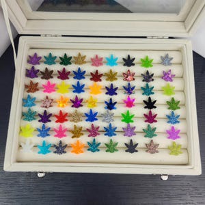 May include: A collection of colorful, glittery marijuana leaf-shaped earrings displayed in a white jewelry box. The earrings come in various colors, including green, blue, purple, and gold, and are arranged neatly on a ring holder.