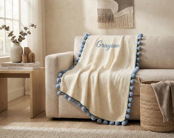 Può includere: Una coperta in maglia color crema con un bordo di pon pon blu lungo i lati e il nome Grayson ricamato in corsivo blu sulla parte superiore del tessuto.