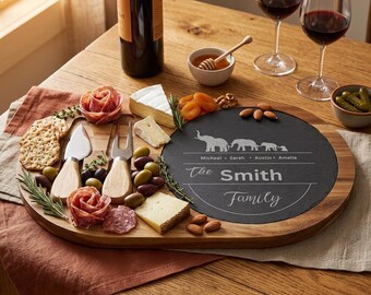 Può includere: Un tagliere in legno con inserto in ardesia, con incisa la scritta "The Smith Family" e sagome di elefanti. Il tagliere contiene formaggio, olive, cracker e salumi, con una bottiglia di vino e due bicchieri.