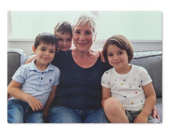 Può includere: Una donna anziana e tre bambini piccoli sono seduti insieme su un divano grigio. La donna e i bambini sorridono alla fotocamera. I bambini indossano abiti casual, tra cui jeans e magliette.