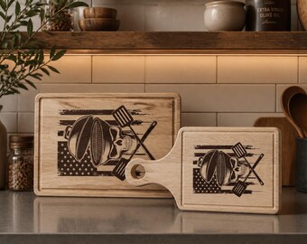 Può includere: Due taglieri in legno con un design a tema barbecue. Il design presenta un teschio, una griglia e spatole su una bandiera americana. Una tavola è rettangolare, l'altra ha un manico.