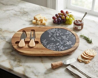 Può includere: Un tagliere ovale in legno con un inserto circolare in ardesia nera decorato da un motivo floreale bianco. Il tagliere presenta due alloggiamenti per un coltello e una forchetta da formaggio con manici in legno, oltre a una spatola con manico in sughero.