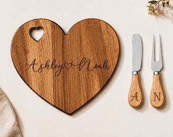 Può includere: Un tagliere in legno a forma di cuore con un foro a forma di cuore, inciso con il testo Ashley e Noah separati da un simbolo a cuore. Accanto si trovano un coltello da formaggio e una forchetta a due rebbi in metallo con manici in legno incisi con A e N.