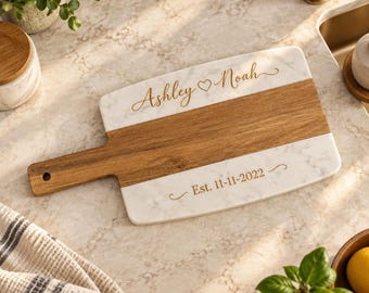 Può includere: Un tagliere rettangolare con un design in marmo bianco e legno marrone dotato di manico. Il tagliere è inciso con il testo Ashley Noah separato da un simbolo a cuore e il testo Est. 11-11-2022 in un carattere corsivo color oro.