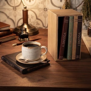 May include: A light-colored wooden box containing four books labeled Volume I, Volume II, Volume III, and Volume V, positioned next to a speckled white ceramic coffee mug on a saucer with a silver spoon, and a black book labeled Volume V.