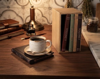 Può includere: Una scatola in legno chiaro contenente quattro libri etichettati Volume I, Volume II, Volume III e Volume V, posizionata accanto a una tazza da caffè in ceramica bianca maculata su un piattino con un cucchiaino d'argento e un libro nero Volume V.