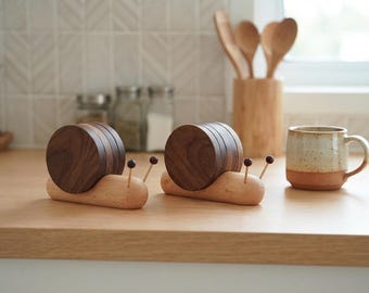 Può includere: Due accessori da cucina in legno a forma di lumaca, caratterizzati da un corpo in legno chiaro e un guscio circolare in legno scuro. Ogni lumaca presenta due sottili antenne sormontate da piccole sfere scure in legno levigato.
