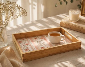 Può includere: Un vassoio da portata rettangolare in bambù con una base a righe verticali rosa e bianche decorata con piccoli motivi floreali. Il vassoio è dotato di manici laterali integrati e contiene una tazza in ceramica bianca riempita di tè.