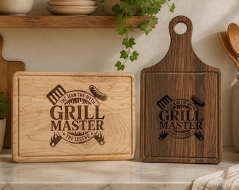 Può includere: Due taglieri in legno con la scritta "GRILL MASTER" e grafica correlata. Un tagliere è di colore chiaro, rettangolare, l'altro è di colore scuro, con un manico. Arredamento cucina.