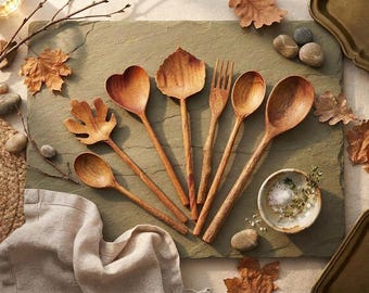 Può includere: Un set di sei utensili da cucina in legno, inclusi cucchiai, una forchetta e un servitore a forma di foglia, disposti su una superficie di pietra piatta accanto a una piccola ciotola in ceramica contenente sale ed erbe, decorato con foglie autunnali essiccate e ciottoli.