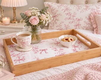 Può includere: Un vassoio da portata rettangolare in bambù con base bianca e un motivo ripetuto di fiori e nastri rosa. Il vassoio è dotato di manici laterali integrati e contiene una tazza in ceramica, una piccola ciotola smerlata e un vaso di fiori in vetro.