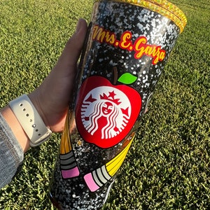 May include: A black and white tumbler with glitter, a red apple with a white Starbucks logo, and yellow pencils. The tumbler has a gold rim and the text "Mrs. E. Garza" on the side.