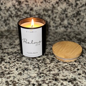 Puede incluir: Un tarro de vela de vidrio negro con una etiqueta blanca que dice "JC Candle Co. Psalms 23:4 Vanilla Birch Hand poured soy wax 7oz". La vela está encendida y la tapa es de madera.
