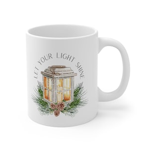May include: White ceramic mug with a design of a lantern with a lit candle inside, surrounded by pine branches and pine cones. The text "Let your light shine" is written in a circle around the lantern.