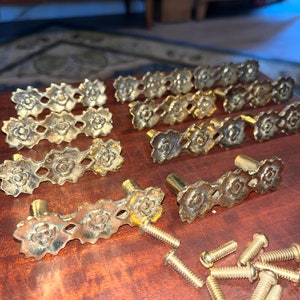 May include: A collection of antique brass drawer pulls with a floral design. The pulls are arranged on a wooden surface, along with several brass screws. The drawer pulls have a vintage aesthetic.
