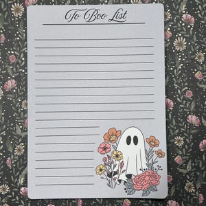 May include: A gray to-do list notepad with the text "To Boo List" at the top. A ghost with two eyes is drawn in the bottom right corner of the notepad, surrounded by flowers in shades of orange, yellow, and pink.