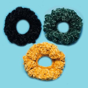 May include: Three plush scrunchies in black, forest green, and golden yellow. These soft, textured hair accessories are arranged on a light blue background. The scrunchies have a fluffy, velvet-like appearance.