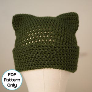 May include: A dark olive green crocheted beanie featuring two pointed cat-ear shapes at the top. The hat has a wide, dense brim and a more open, airy stitch pattern on the crown. A circular white badge in the corner displays the text PDF Pattern Only.