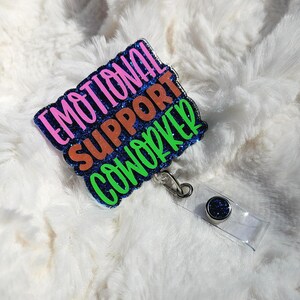 May include: A rectangular badge with the words "EMOTIONAL SUPPORT COWORKER" in pink, orange, and green glitter lettering. The badge has a blue glitter border and a clear plastic retractable reel with a silver clip.