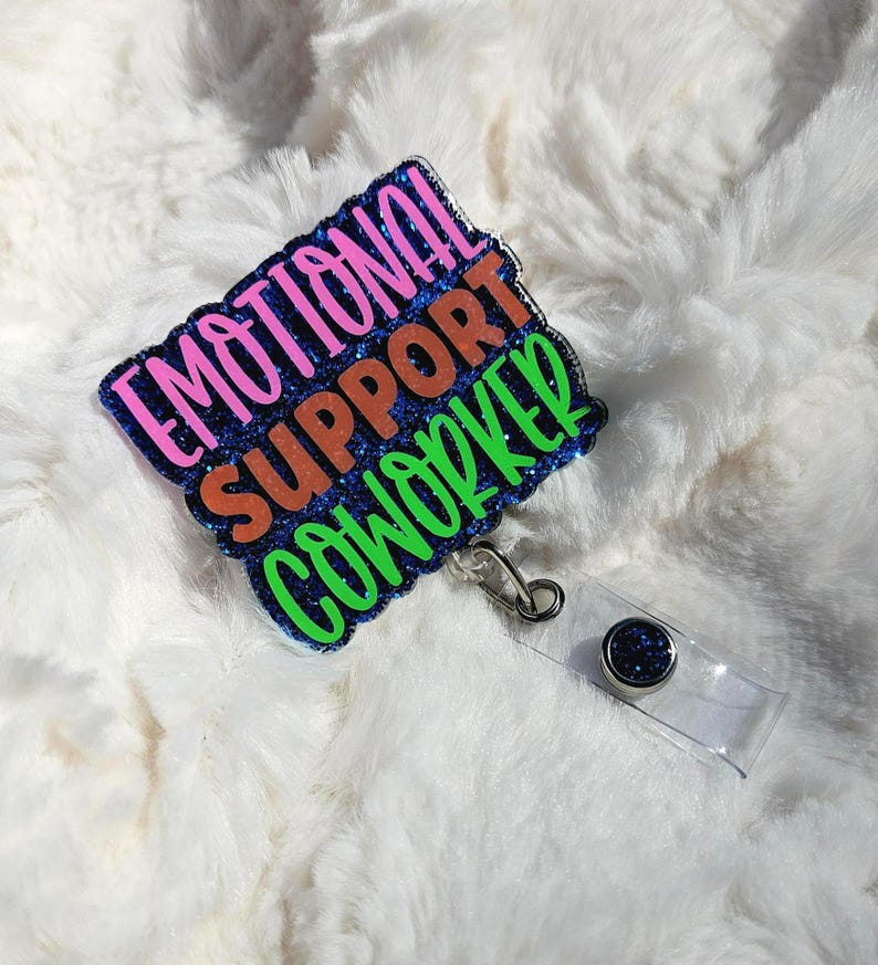 May include: A rectangular badge with the words "EMOTIONAL SUPPORT COWORKER" in pink, orange, and green glitter lettering. The badge has a blue glitter border and a clear plastic retractable reel with a silver clip.
