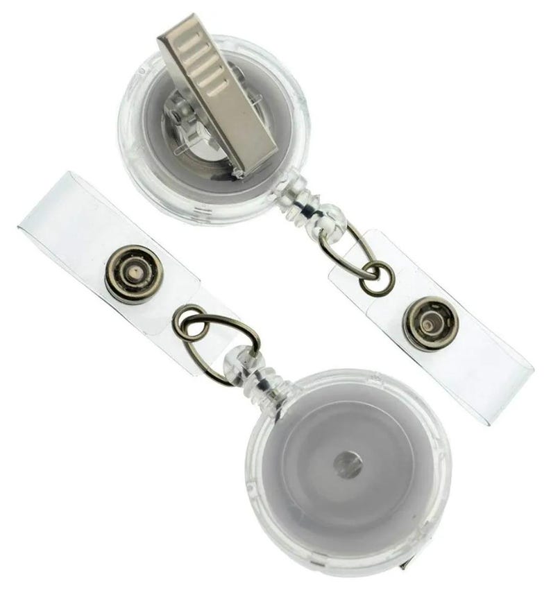 May include: Two clear plastic retractable badge holders with metal clips and clear straps. The badge reels have a circular design with a silver-coloured metal clip on top and a clear strap with a metal snap closure.