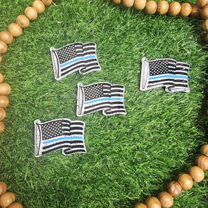 May include: Four small flag pins featuring a black and white American flag design with a blue stripe. The pins are displayed on green grass, with a wooden bead necklace in the background.