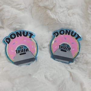 May include: Two donut-shaped pins with pink icing and colourful sprinkles. The pins have a blue glitter border and the word "DONUT" at the top. Below the donut is the text "OF TRUTH" and a grey object.