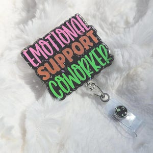 May include: A rectangular badge reel displaying the text "EMOTIONAL SUPPORT COWORKER" in pink, orange, and green. The badge has a black background with glitter and a clear plastic reel with a metal clip.