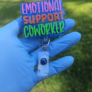 May include: A badge reel with the words "EMOTIONAL SUPPORT COWORKER" in pink, orange, and green glitter lettering on a blue glitter background. The reel has a clear plastic holder with a blue glitter accent.