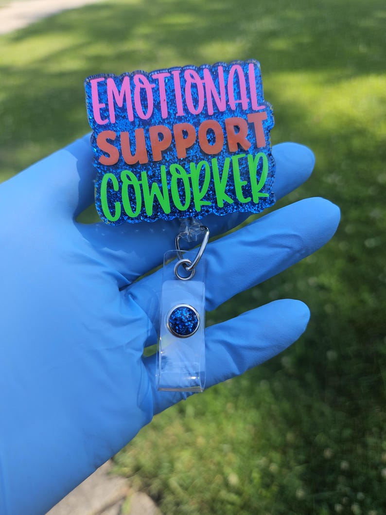 May include: A badge reel with the words "EMOTIONAL SUPPORT COWORKER" in pink, orange, and green glitter lettering on a blue glitter background. The reel has a clear plastic holder with a blue glitter accent.