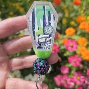 May include: A coffin-shaped badge reel with a green and white striped background, green slime, and a cartoon character. The reel has a green top, a beaded purple and black accent, and a clear clip.