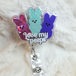 May include: A badge reel with three glittery Peeps marshmallow chicks in purple, teal, and pink. The text "Love my peeps" is written in cursive. The reel has a clear plastic strap and a metal clip.