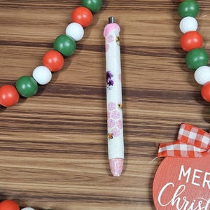 May include: A white pen with a pink top and a glittery finish. The pen has a design of bees and flowers. The pen is sitting on a wooden surface with a red and white Christmas ornament in the background.