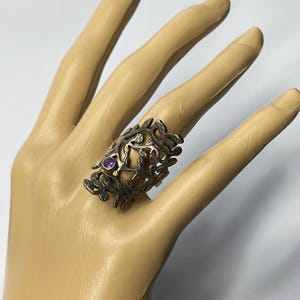May include: A wide, dark-toned metal ring featuring an intricate, openwork branch or vine pattern. The band is accented with a single, small, round, purple gemstone set into the textured metal surface, creating a detailed and organic appearance.