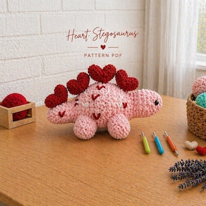 Puede incluir: Un estegosaurio rosa hecho a mano con detalles de corazón rojo. El dinosaurio tiene espinas en forma de corazón rojo y pequeños corazones rojos en su cuerpo. La imagen también incluye ganchos de ganchillo, ovillos de lana y una pequeña planta.