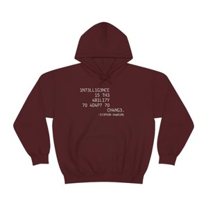 May include: A maroon hoodie with a drawstring hood and a kangaroo pocket. The front of the hoodie features white text that reads "1NT3LL1G3NCE 15 TH3 4B1L17Y 70 4D4P7 70 CH4NG3. -573PH3N H4WK1NG."