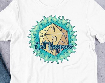 Puede incluir: Camiseta blanca con un diseño de dado dorado de múltiples caras, mostrando los números 14 y 20, rodeado por una llama turquesa y azul. El texto "Crit Happens" está escrito debajo. La camiseta se muestra sobre una superficie clara.