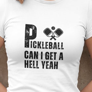 Może przedstawiać: Biała koszulka z czarnym nadrukiem, na którym widnieje napis "Pickleball Can I Get A Hell Yeah" oraz dwie czarne rakiety do pickleballa.