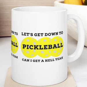 May include: White ceramic coffee mug with a yellow, black, and gray design. The mug features three yellow pickleball paddles with gray holes and the text "Let's get down to pickleball. Can I get a hell yeah."
