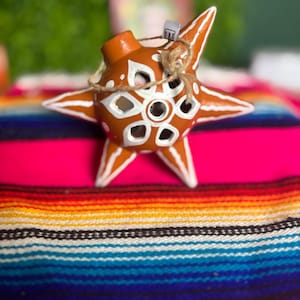 May include: A star-shaped terracotta ornament featuring a central cutout pattern with white painted accents and a brown twine loop for hanging. The surface is a warm reddish-brown clay with white lines tracing the points of the star.