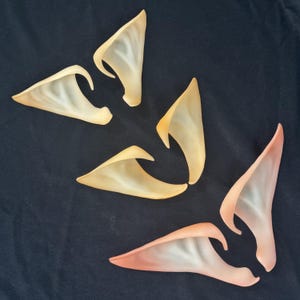 May include: Three pairs of pointed prosthetic ears in varying shades of beige, peach, and light pink. Each pair features a curved, tapered design with a smooth, semi-translucent finish, arranged in a scattered pattern against a dark background.