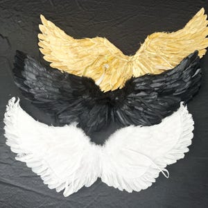 May include: Three sets of decorative wings arranged vertically. The top set is gold, the middle set is black, and the bottom set is white. Each pair of wings is constructed from layered feathers, featuring a wide, arched shape with pointed tips.