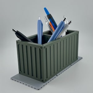 Könnte beinhalten: Ein grauer, rechteckiger Behälter mit einem Wellendesign, der einem Versandcontainer ähnelt, wird als Stifthalter verwendet. Er enthält eine Vielzahl von Stiften, darunter blaue, schwarze und silberne.