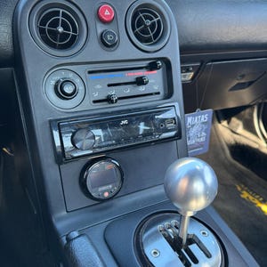 Może przedstawiać: Wnętrze samochodu z czarną deską rozdzielczą. Widoczne są otwory wentylacyjne, radio i elementy sterujące. Widoczna jest srebrna gałka dźwigni zmiany biegów oraz mały cyfrowy wskaźnik. Tekst "MIATAS MOUNTAINS" znajduje się na zawieszce.