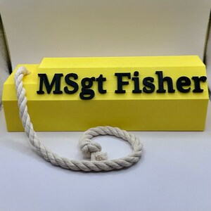 May include: A yellow wooden block with the text "MSgt Fisher" in black lettering. A white rope is attached to the block.